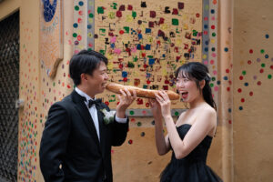 ニースのカラフルな壁の前でフランスパンを頬張る新郎新婦