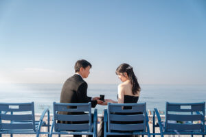 ニースの海辺に並ぶかわいい青いベンチに座って婚約指輪の箱を持つ新郎新婦